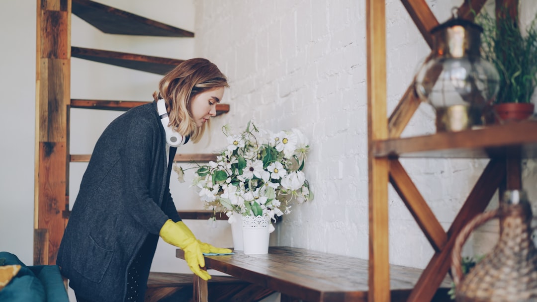 Blond housewife wearing protective rubber gloves is dusting a table at home doing housework. Wireless headphones, staircase and beautiful wooden furniture is visible.